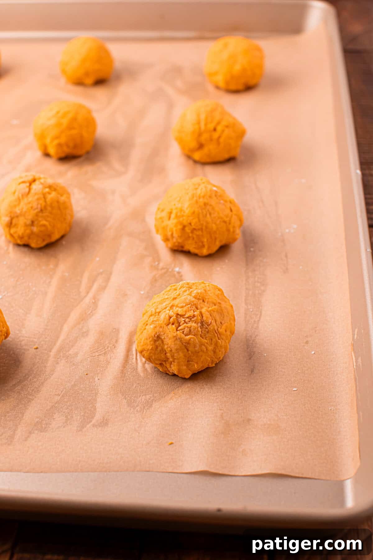 Ten unbaked pumpkin dough balls are neatly arranged and spaced out on a parchment-lined baking sheet, showcasing their light orange hue and slightly textured surface, perfectly prepped for baking.