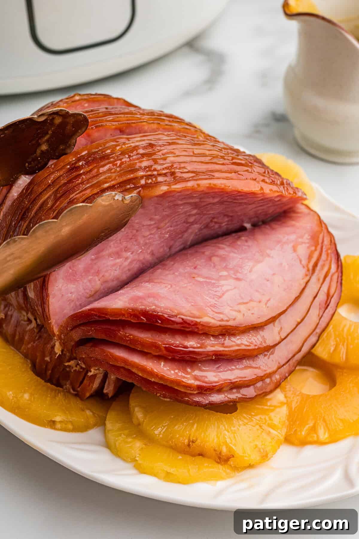 Irresistible Slow Cooker Ham with Pineapple Brown Sugar Glaze 2 A glistening platter of boneless spiral sliced ham, generously glazed and surrounded by vibrant pineapple rings. A pair of metal tongs gently lifts a perfect slice of ham, showcasing its juicy texture. A black slow cooker, hinting at the ease of preparation, sits softly blurred in the background.