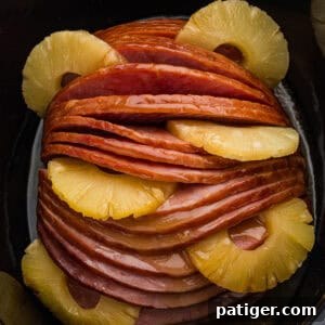 Irresistible Slow Cooker Ham with Pineapple Brown Sugar Glaze 12 A glazed spiral sliced ham sits in a black slow cooker with pineapple rings arranged between the slices and around the edges.