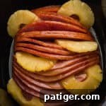 Irresistible Slow Cooker Ham with Pineapple Brown Sugar Glaze 13 A glazed spiral sliced ham sits in a black slow cooker with pineapple rings arranged between the slices and around the edges.