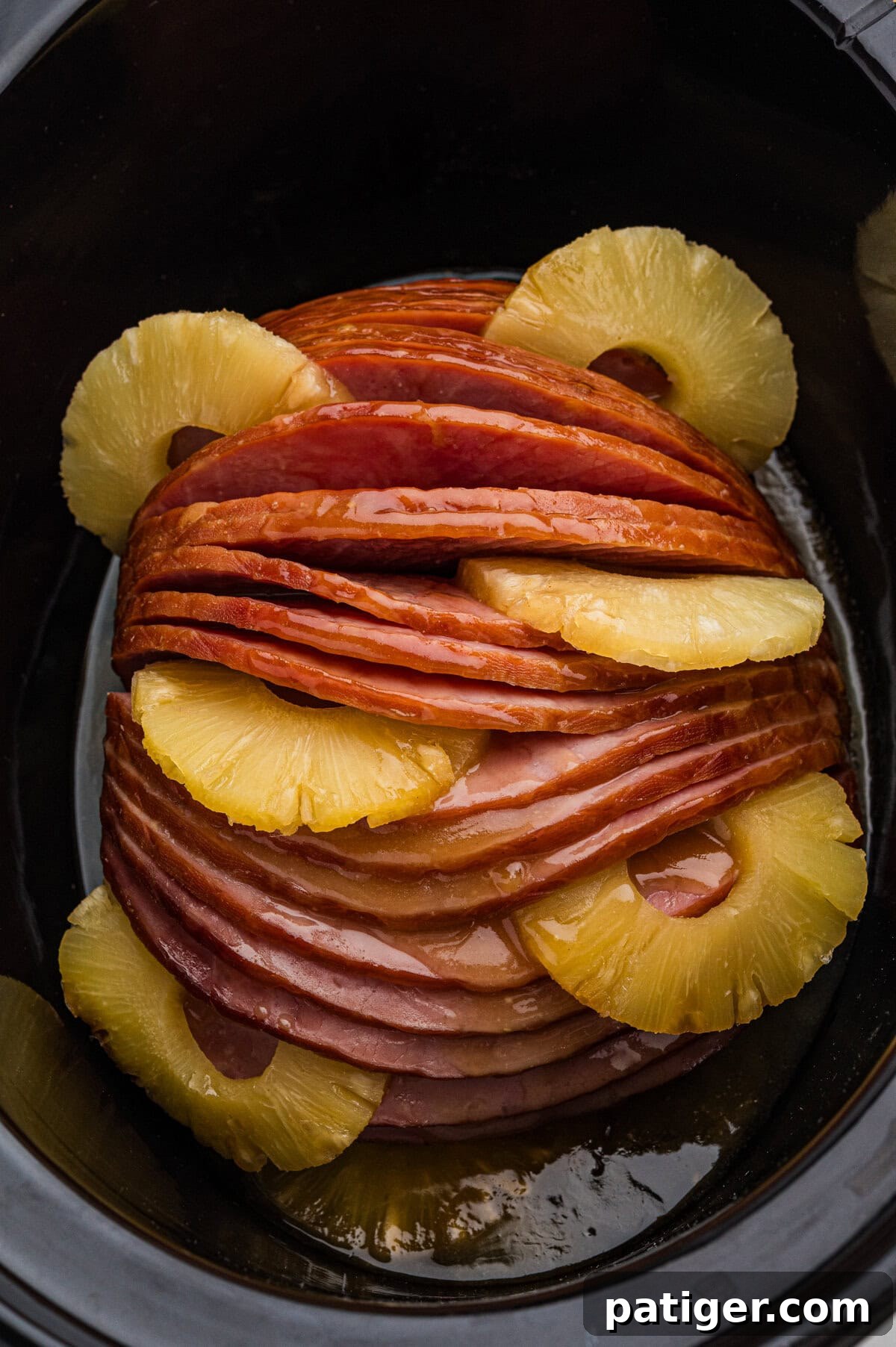 Irresistible Slow Cooker Ham with Pineapple Brown Sugar Glaze 3 A beautifully glazed spiral sliced ham nestled within a black slow cooker, with vibrant pineapple rings artfully arranged between its slices and around the edges, ready for cooking.