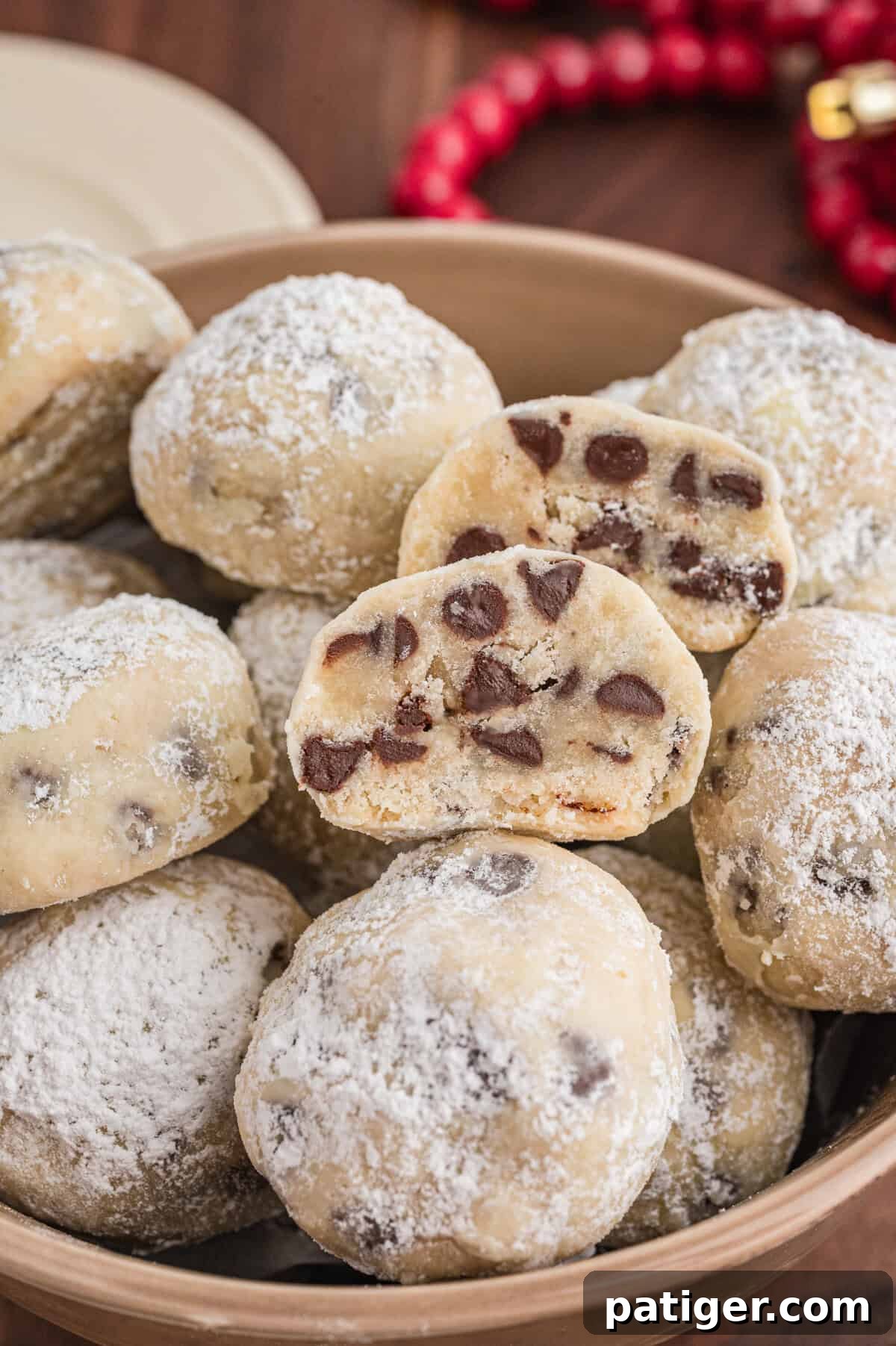 Decadent Chocolate Chip Snowball Bites 2 Chocolate chip snowball cookies in a bowl. The cookies are coated in powdered sugar, and a halved cookie on top shows the interior full of mini chocolate chips.