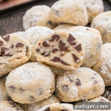 Mini chocolate chip snowball cookies stacked on a tray, with one cut open to show the chocolate chips inside. The cookies are coated in powdered sugar.