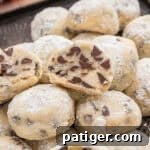Mini chocolate chip snowball cookies stacked on a tray, with one cut open to show the chocolate chips inside. The cookies are coated in powdered sugar.