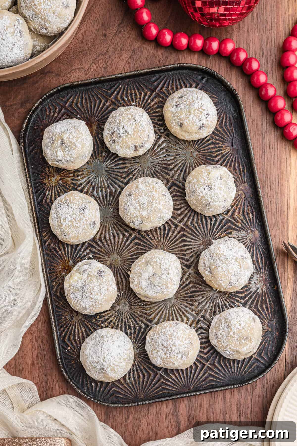 Decadent Chocolate Chip Snowball Bites 3 Snowball cookies with chocolate chips coated in confectioners sugar on a dark patterned tray arranged in neat rows.