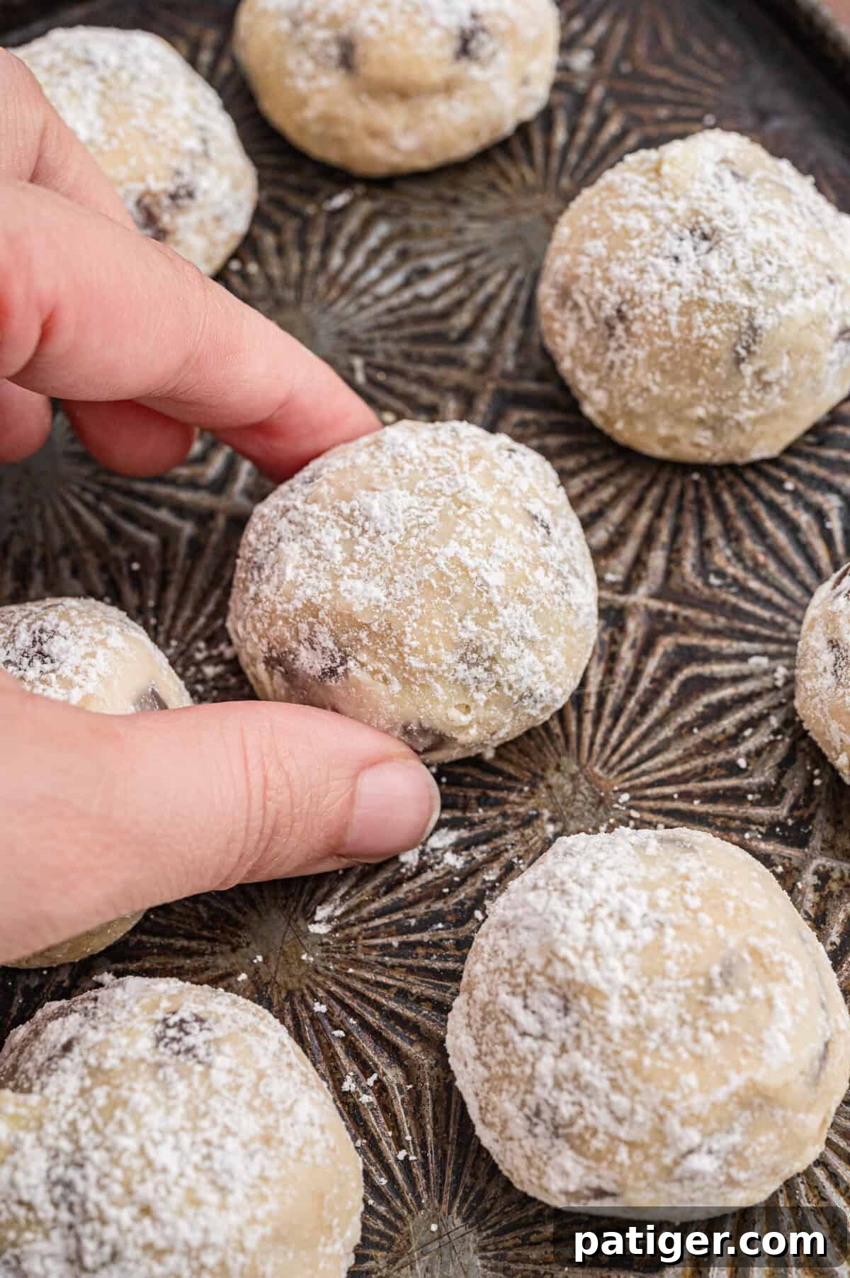 Decadent Chocolate Chip Snowball Bites 9 Close-up of a hand picking up a chocolate chip snowball cookie from a dark, patterned tray. The cookie is round, golden, and covered in powdered sugar. Several more cookies are visible on the tray.