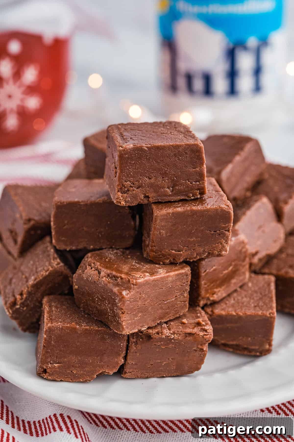 A stack of marshmallow fluff chocolate fudge pieces on a white plate placed on a red-striped cloth. In the background are festive decorations, including a red mug and a jar of Marshmallow Fluff.