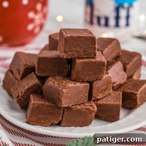 A stack of marshmallow fluff chocolate fudge pieces on a white plate placed on a red-striped cloth. In the background are festive decorations, including a red mug and a jar of Marshmallow Fluff.