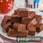 A stack of marshmallow fluff chocolate fudge pieces on a white plate placed on a red-striped cloth. In the background are festive decorations, including a red mug and a jar of Marshmallow Fluff.