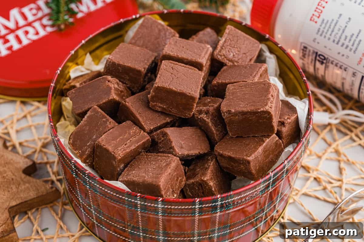 A round red plaid holiday tin filled to the top with square chocolate marshmallow fluff fudge pieces. A jar of Marshmallow Fluff and a red "Merry Christmas" lid in the background.