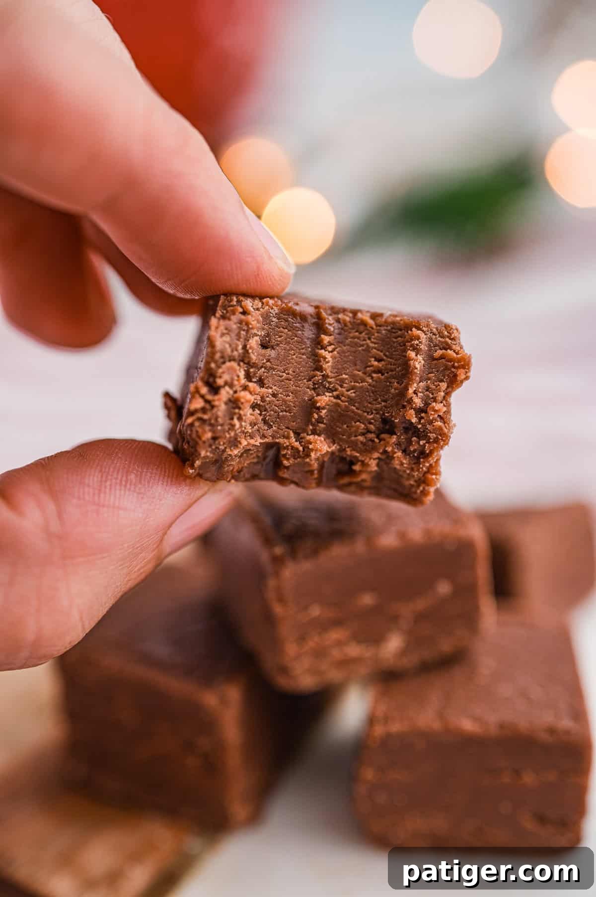 A close-up of a hand holding a square piece of chocolate fudge with a bite taken out, revealing the smooth, creamy interior.