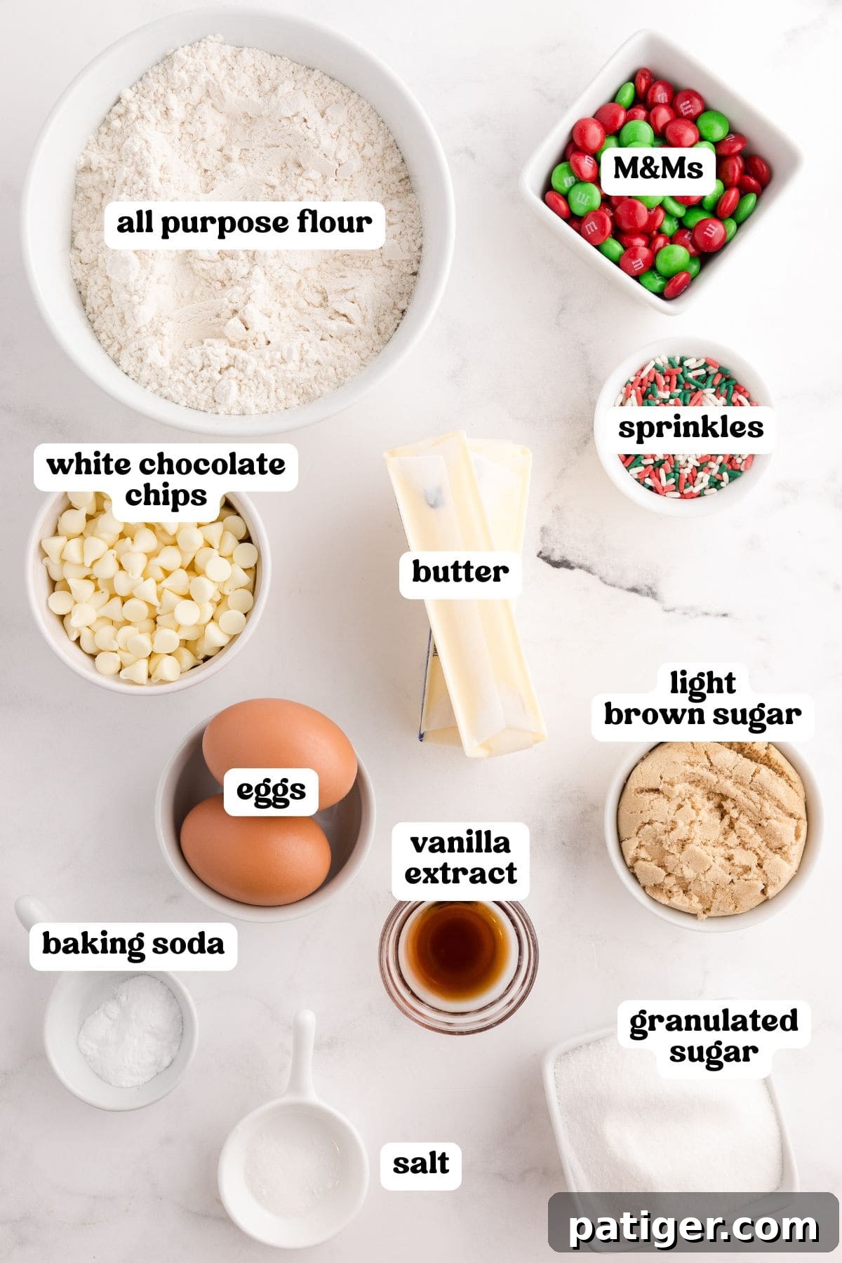 All the Christmas M&M cookie ingredients laid out in separate bowls on a white background: all-purpose flour, M&Ms, white chocolate chips, sprinkles, butter, light brown sugar, granulated sugar, eggs, vanilla extract, baking soda, and salt.