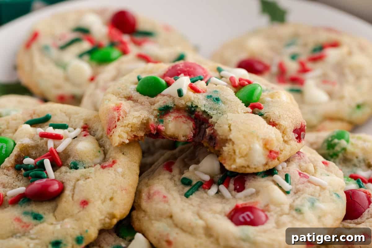 A close-up of a stack of fresh-baked Christmas M&M cookies with white chocolate chips and festive sprinkles, with one cookie having a bite taken out of it, revealing its soft interior.