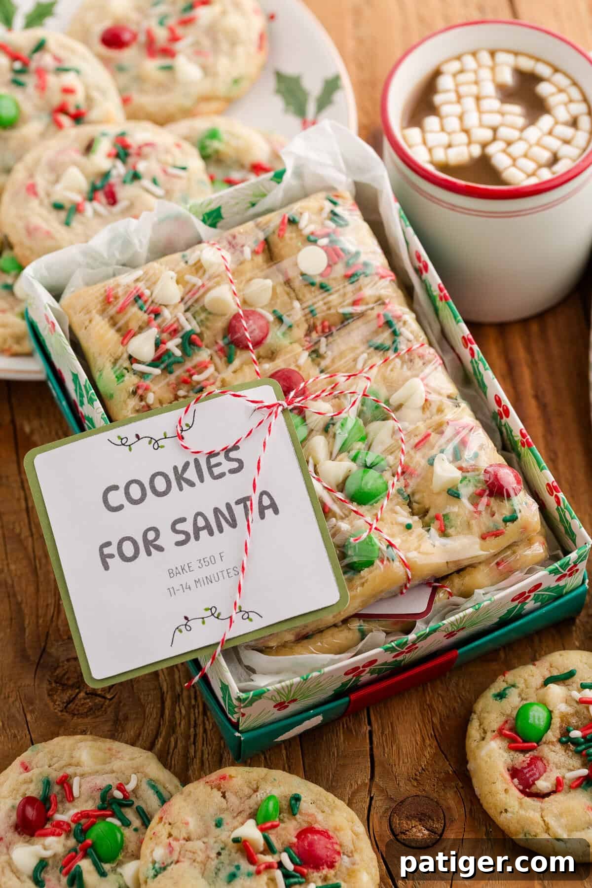 A beautifully arranged holiday gift box filled with rectangular cookie dough portions, adorned with red and green M&Ms, white chocolate chips, and festive sprinkles. Each portion is carefully wrapped in plastic and tied with charming red and white string, accompanied by a label that proudly reads “COOKIES FOR SANTA – BAKE 350 F, 11–14 MINUTES.” The gift box is surrounded by baked cookies, a festive holly-decorated plate, and a cozy white mug of hot cocoa topped with mini marshmallows, evoking a warm and inviting holiday scene.