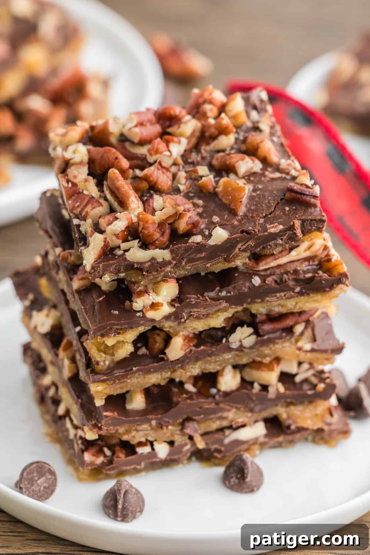 A stack of chocolate and pecan-topped saltine cracker toffee pieces on a white plate. Chocolate chips are scattered around the plate.