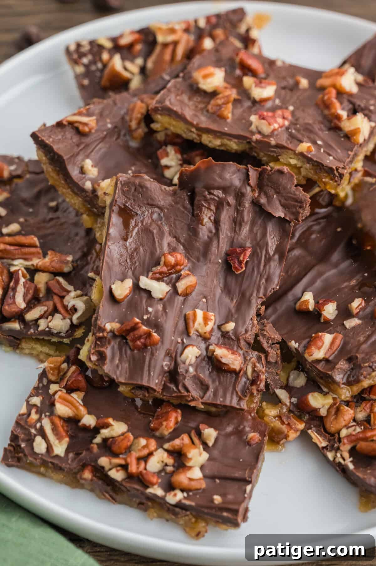 A white plate filled with overlapping pieces of saltine cracker toffee. The toffee is coated in chocolate and topped with scattered chopped pecans.