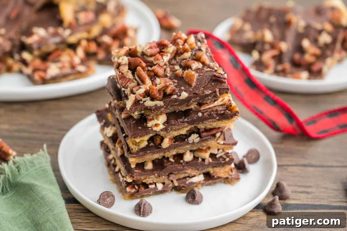 A stack of saltine cracker toffee topped with chopped pecans and layered with chocolate sits on a white plate. Chocolate chips are scattered around the plate, with blurred toffee pieces in the background.