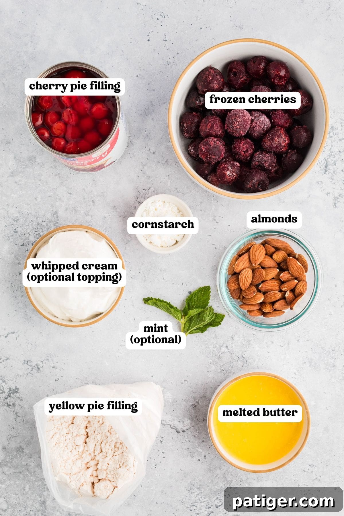 Ingredients for cherry cobbler displayed on a gray surface, each labeled: cherry pie filling, frozen cherries, cornstarch, almonds, whipped cream (optional topping), mint (optional), yellow pie filling mix, and melted butter.