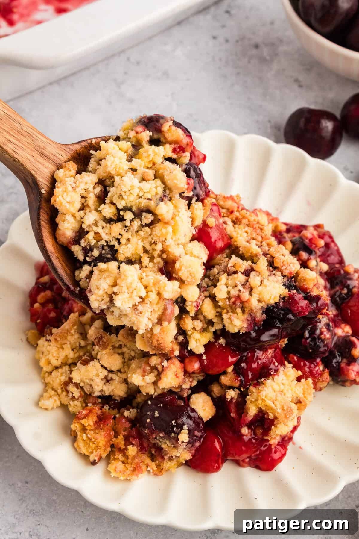 A wooden spoon scooping cherry cobbler onto a white scalloped plate. The cobbler has a crumbly topping with chunks of yellow cake mix and cherries visible in the filling.