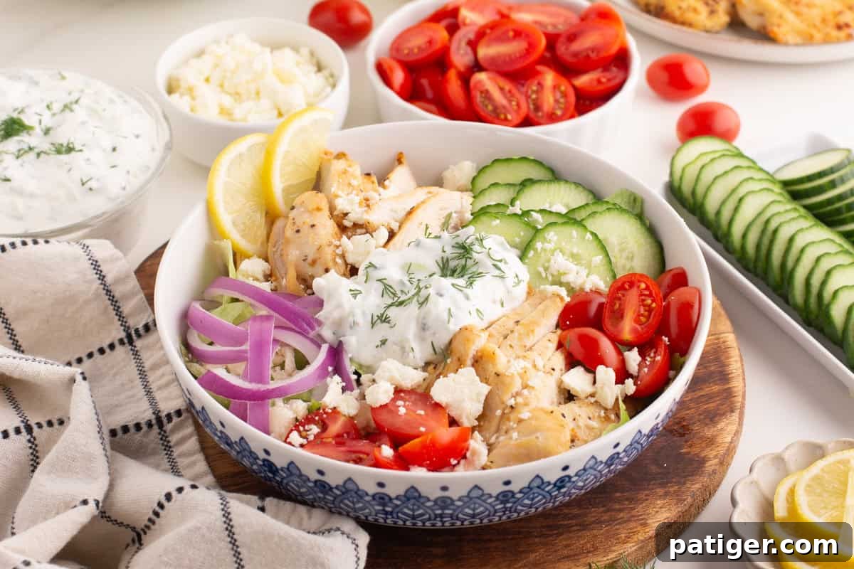 A vibrant chicken gyro bowl featuring grilled chicken slices, halved cherry tomatoes, fresh cucumber slices, thinly sliced red onion, crumbled feta, and a generous dollop of creamy tzatziki sauce, garnished with fresh dill. The bowl rests on a rustic wooden board, surrounded by small bowls of individual ingredients and a striped linen towel.