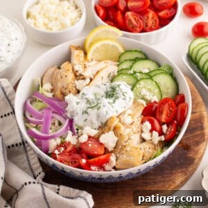 Chicken gyro bowl with grilled chicken, sliced cucumbers, cherry tomatoes, red onions, tzatziki sauce with dill, crumbled feta, and lemon slices.