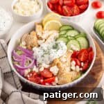 Chicken gyro bowl with grilled chicken, sliced cucumbers, cherry tomatoes, red onions, tzatziki sauce with dill, crumbled feta, and lemon slices.