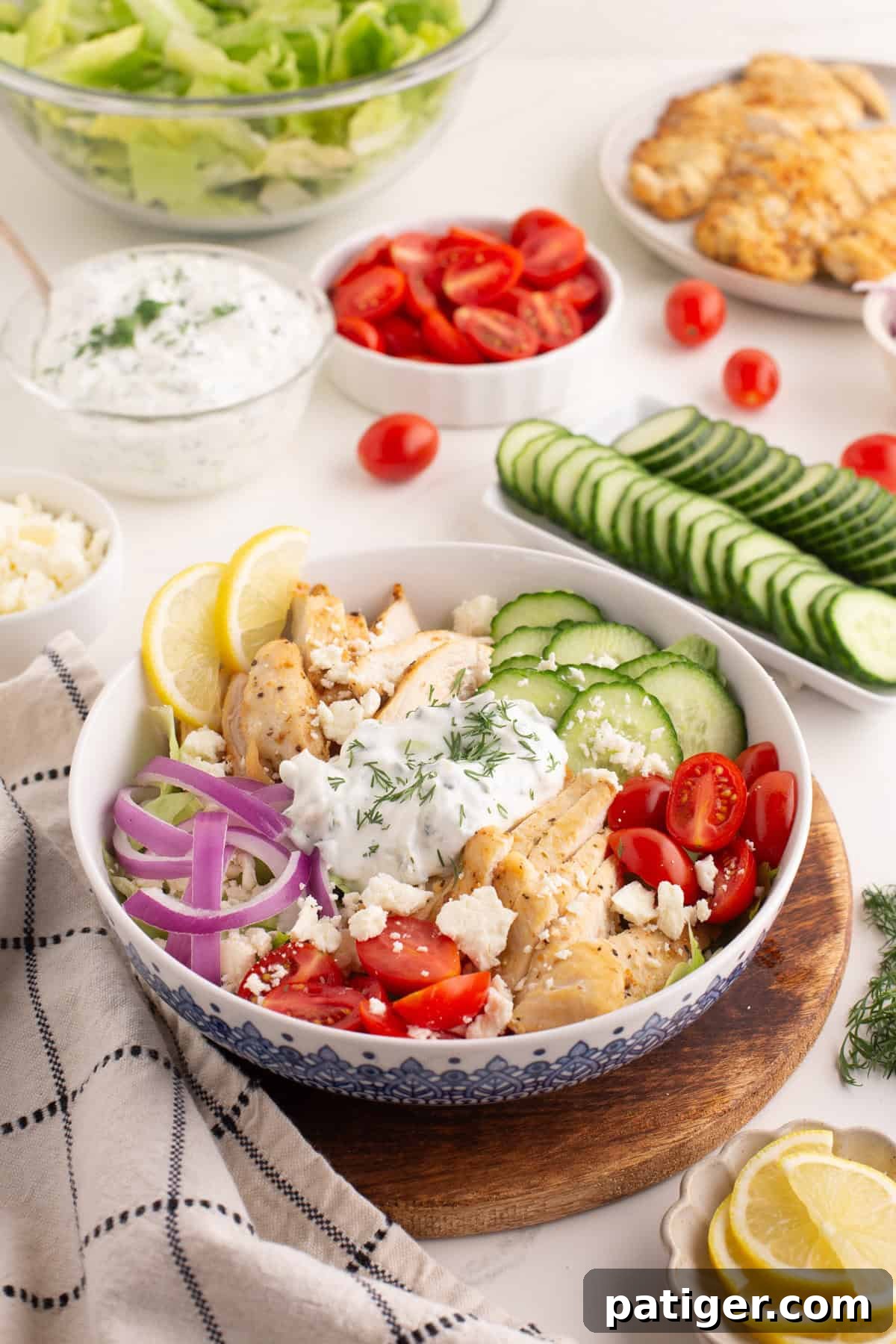 A beautifully presented chicken gyro bowl, brimming with grilled chicken, crisp cucumber slices, juicy cherry tomatoes, thinly sliced red onions, a generous swirl of tzatziki sauce adorned with dill, crumbled feta cheese, and bright lemon slices. The background shows various bowls containing prepped ingredients like lettuce, tomatoes, cucumber, feta, and cooked chicken, emphasizing its meal prep potential.