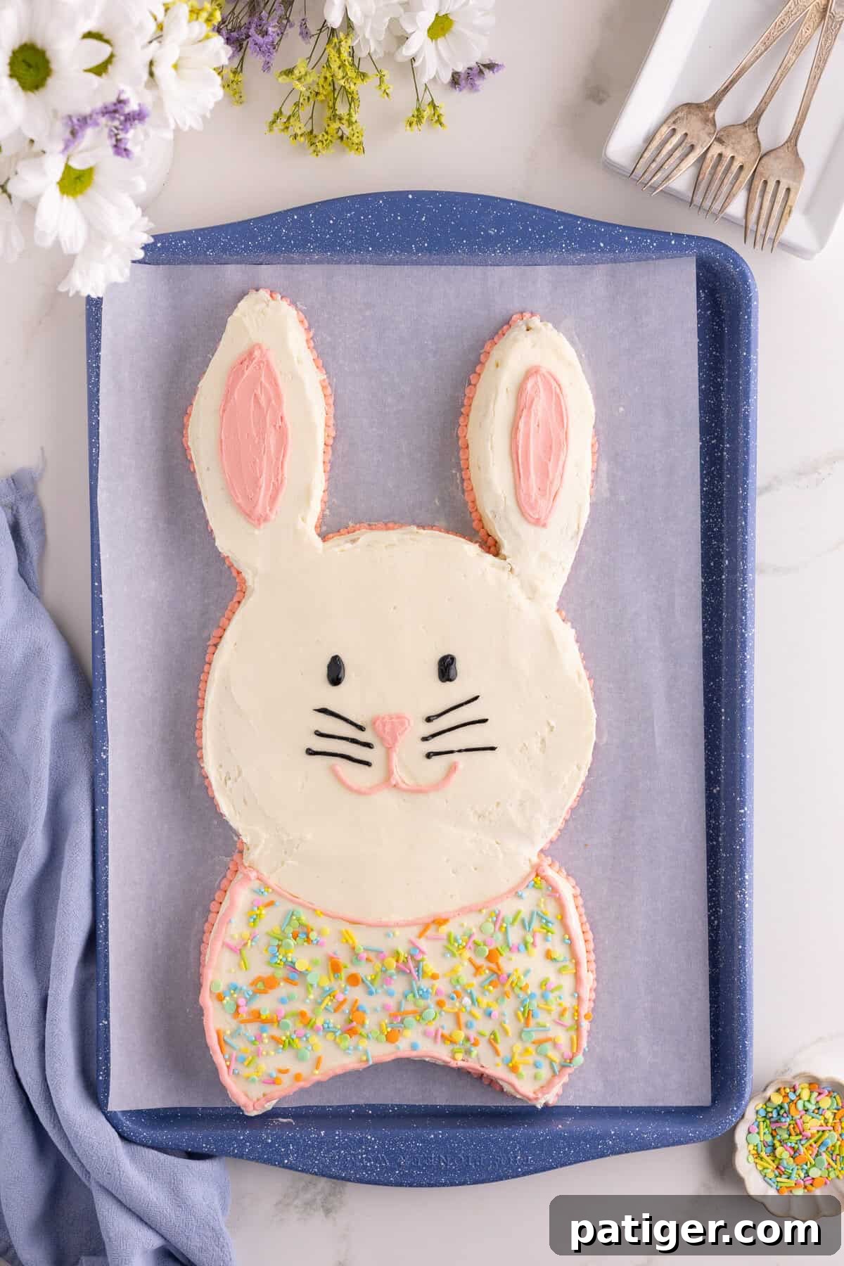 Charming Easter Bunny Cake 2 Overhead view of an Easter bunny cake frosted in white with pink ear details and piped facial features. Adorned with pastel sprinkles.