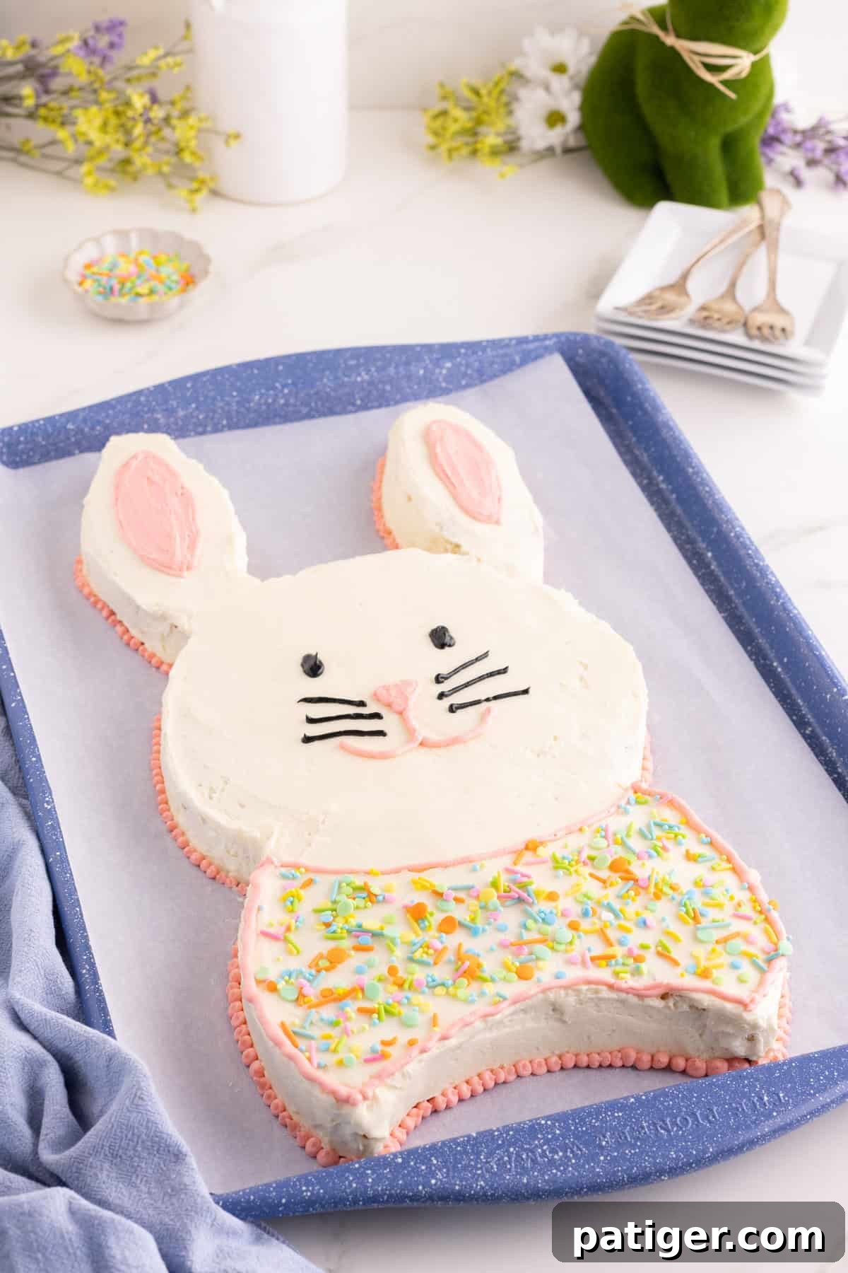 Charming Easter Bunny Cake 5 Bunny-shaped cake with white frosting and pink inner ears, black eyes and whiskers, and a pink nose and mouth. The lower half is decorated like a bow tie with pastel sprinkles and outlined in pink piping.