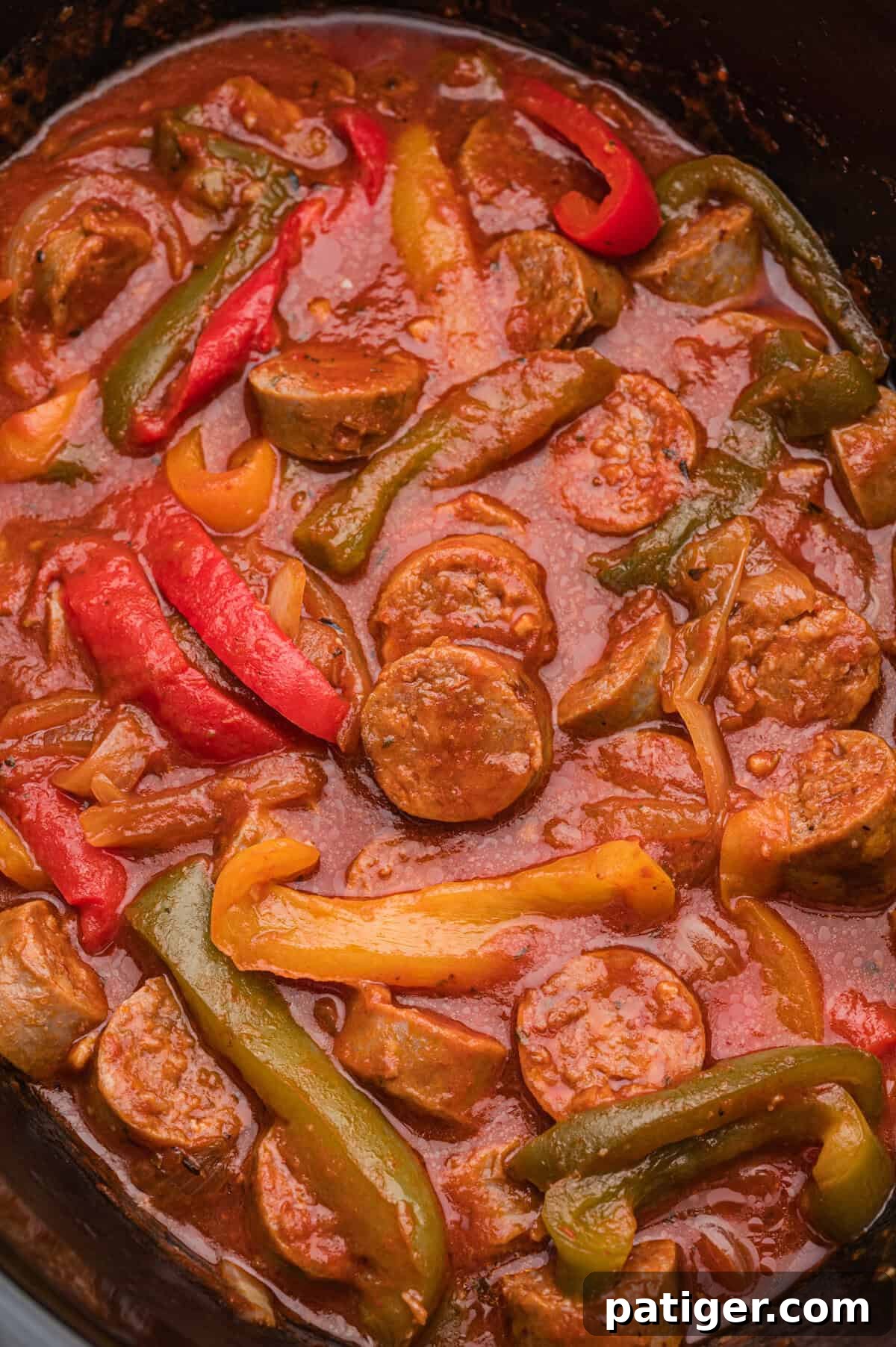 A close-up view of a cooked sausage and peppers dish in a slow cooker, showing sliced sausage rounds, red, yellow, and green bell pepper strips, and onions coated in thick marinara sauce.
