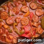 Overhead image showing sliced sausage, green, red, and yellow bell peppers, and onions simmered in red marinara sauce inside a crockpot.