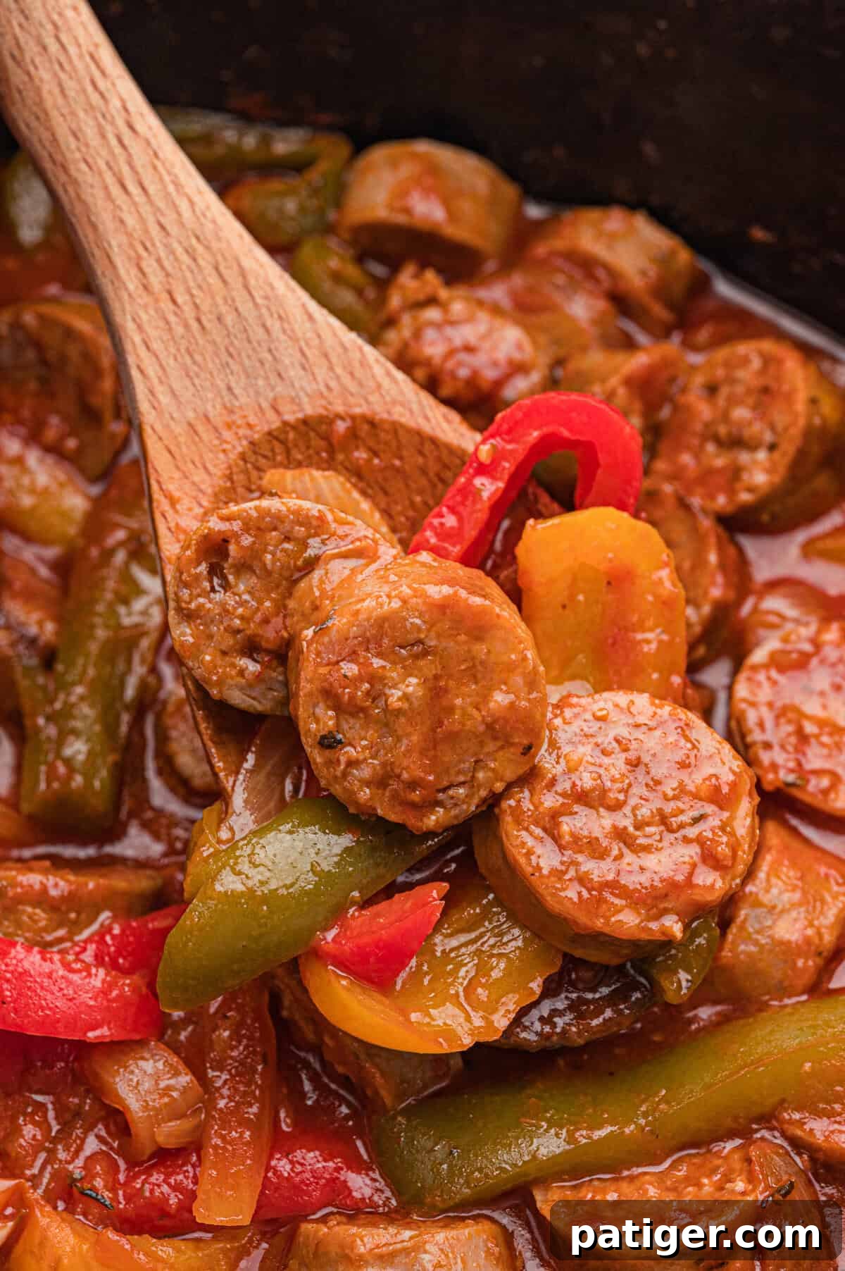 A wooden spoon holds sliced sausage, red and yellow bell pepper strips, and onion over a crockpot of sausage and peppers in marinara sauce.