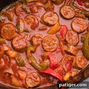 Overhead image showing sliced sausage, green, red, and yellow bell peppers, and onions simmered in red marinara sauce inside a crockpot.