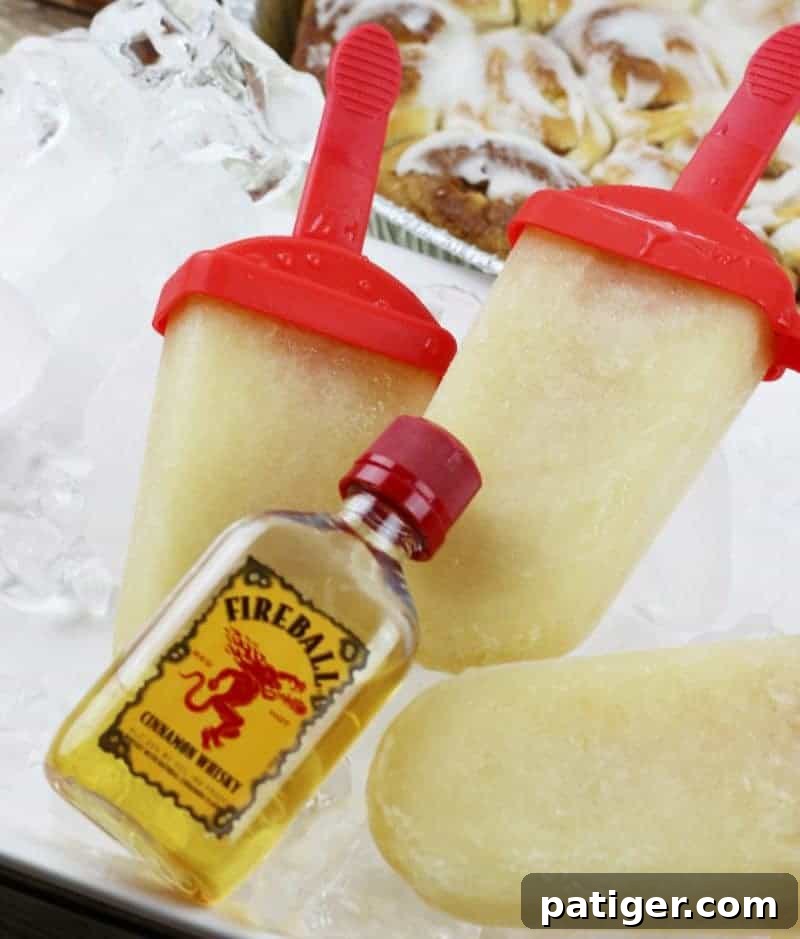 Fireball Whiskey Cinnamon Roll Ice Pops arranged on a bed of ice, with freshly baked cinnamon buns and an airplane-sized bottle of Fireball whiskey in the background. This image showcases the delicious, cinnamon-infused adult ice pops.