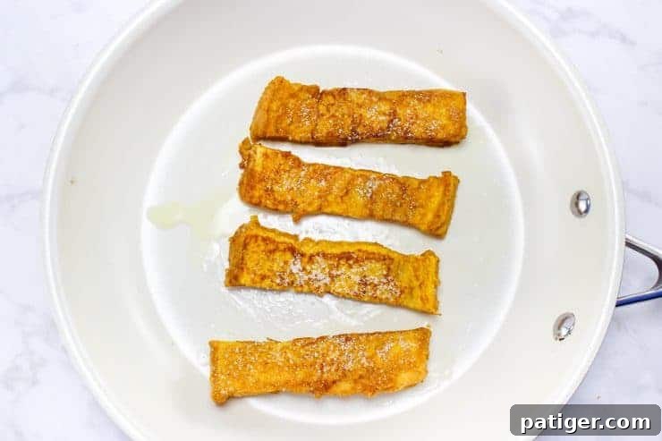 Four golden brown French toast sticks in frying pan, cooked to perfection.