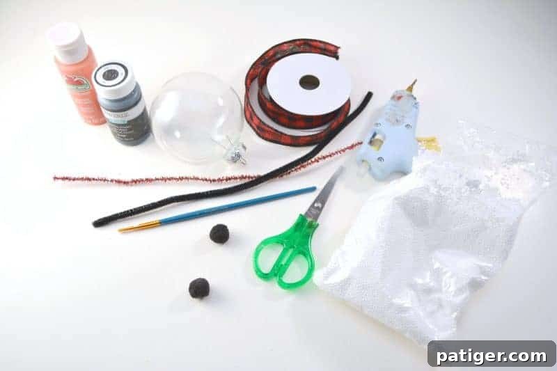 A flat lay of snowman ornament craft supplies including clear plastic ball fillable ornament, small white styrofoam balls, orange and black craft paint, red and black ribbon, scissors, pipe cleaners, pom poms, mini glue gun, and a small paint brush.