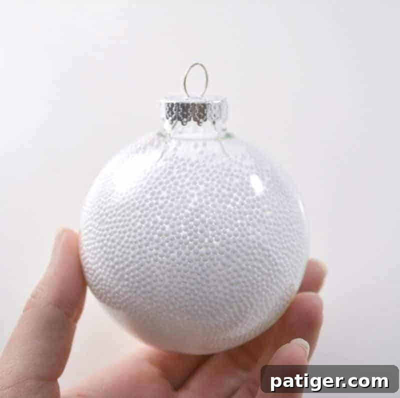 Clear plastic bulb ornament being filled with tiny white fake snow balls, creating a frosty, wintry base for a handmade snowman Christmas ornament.