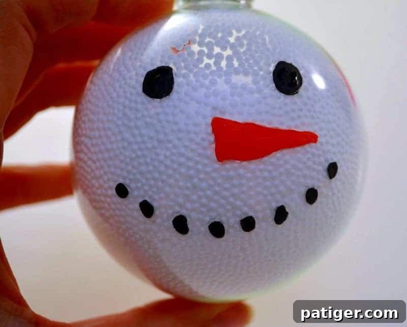 A snowman ornament in progress, showing a cheerful face painted with black circles for eyes, an array of small black dots for a coal mouth, and a vibrant orange triangle for the carrot nose.