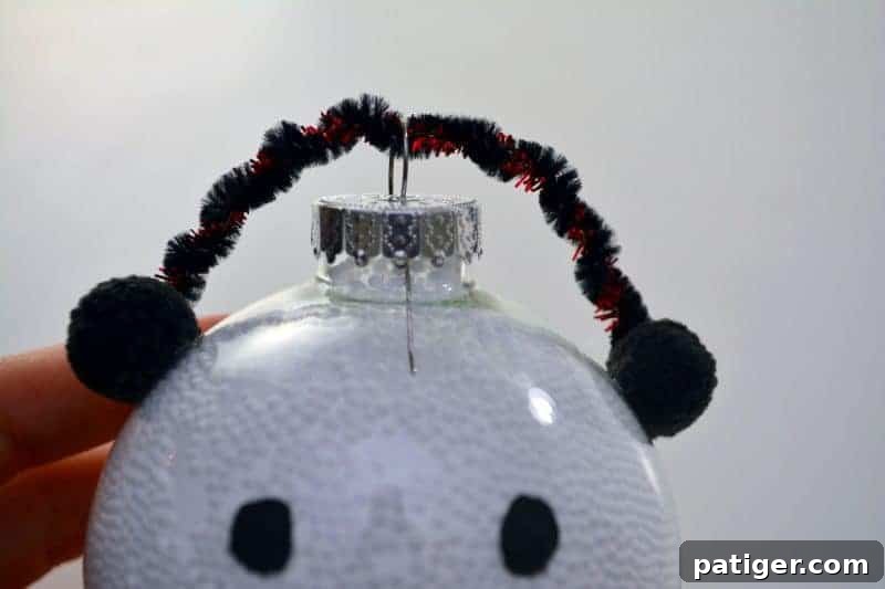 Close-up of a handmade snowman ornament showing colorful pom-poms glued on either side of the clear ball, connected by twisted pipe cleaners forming the earmuffs, adding a whimsical touch.