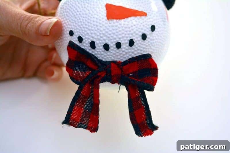 A charming red and black checkered ribbon bow glued underneath the painted face of a handmade snowman ornament, adding a festive and decorative touch to the DIY Christmas decoration.