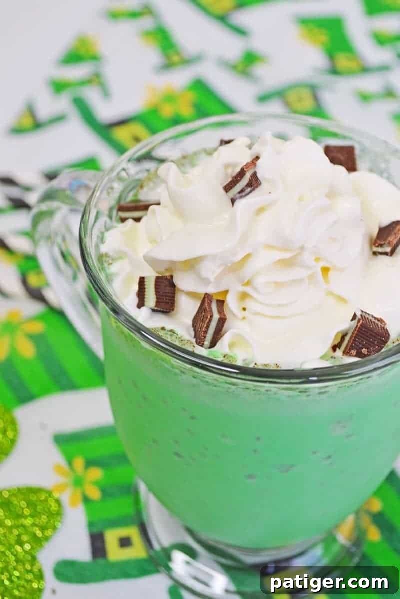 A close-up view of a homemade Shamrock Shake in a glass mug, featuring a rich, bright green color topped with a generous swirl of whipped cream, finely chopped Andes Mints, and a stylish white and black striped paper straw. The mug is placed on a festive leprechaun hat patterned tablecloth, emphasizing its St. Patrick's Day theme.