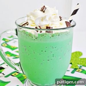 A vibrant green mint chocolate chip milkshake, crowned with fluffy whipped cream and a generous sprinkle of chopped Andes Mints, served in a classic glass mug with a white and black striped paper straw. The mug rests on a festive leprechaun hat themed tablecloth.