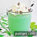 A vibrant green mint chocolate chip milkshake in a glass mug with whipped cream, chopped Andes Mints, and a white and black striped paper straw. The mug rests on a leprechaun hat tablecloth.