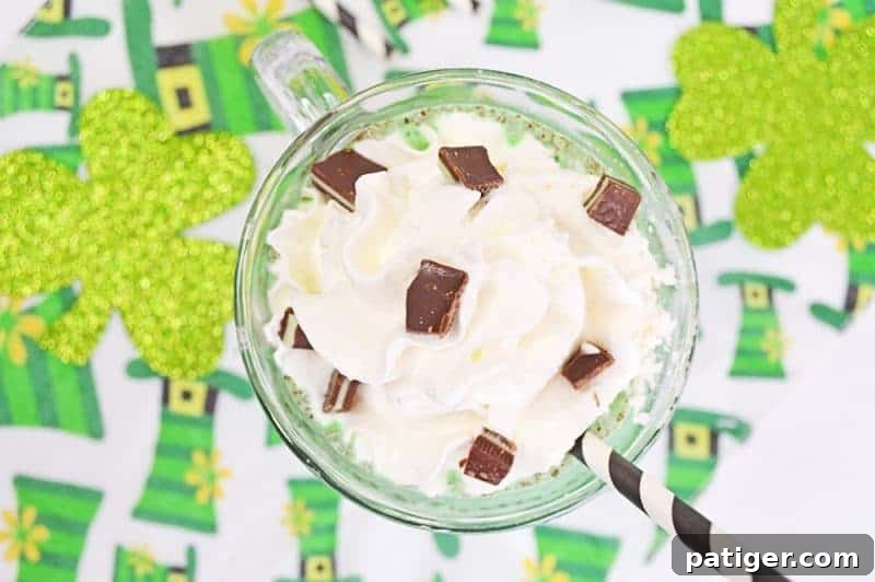 A finished mint chocolate chip milkshake, perfectly blended and topped with a generous swirl of whipped cream and chopped mint chocolate pieces, served in a clear glass with a black and white striped paper straw.