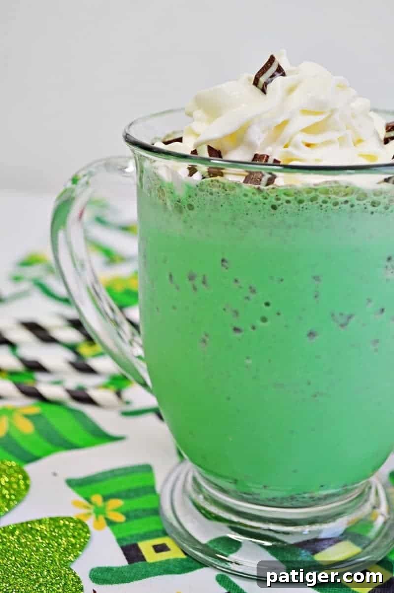 A brightly lit, perfectly crafted mint chocolate chip milkshake in a clear glass mug, topped with a cloud of whipped cream, a sprinkle of chopped Andes Mints, and a white and black striped paper straw. The mug is placed on a green leprechaun hat tablecloth, ready for a St. Patrick's Day celebration.
