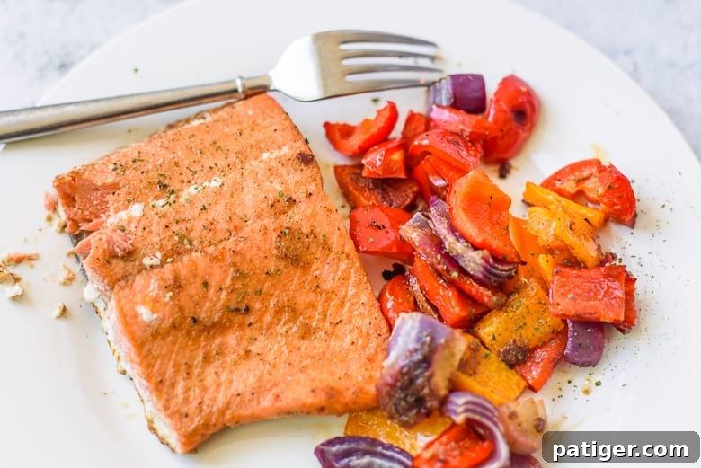 A perfectly cooked salmon fillet, adorned with fresh herbs, resting on a bed of roasted bell peppers and onions, all served on a clean white plate with a fork beside it.