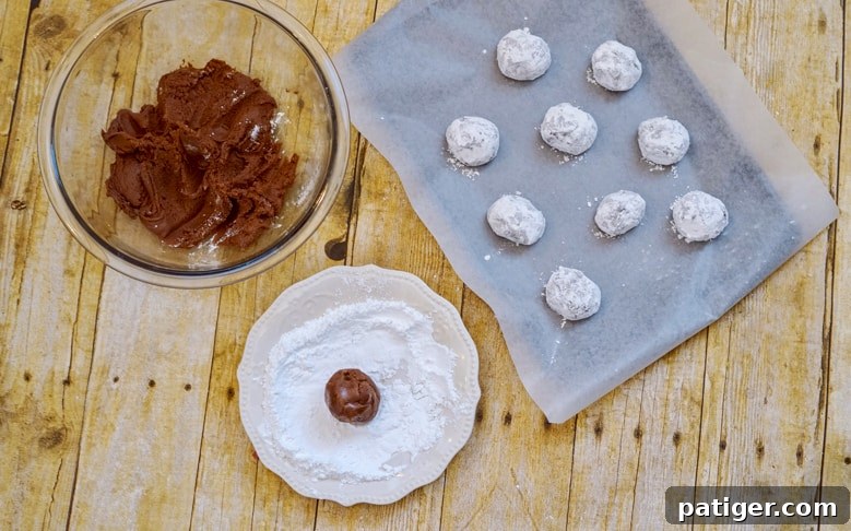 Chocolate fudge cookie dough balls being rolled in powdered sugar and placed on a lined baking sheet, ready for the oven.