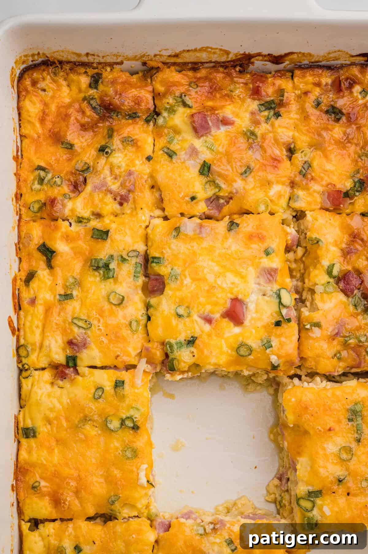 Overhead view of a white baking dish with a sliced breakfast casserole, featuring melted cheese, ham, and green onions, with the center piece removed.