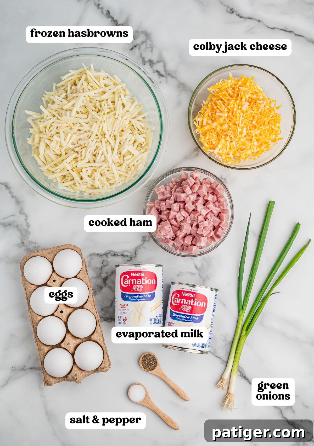 Labeled ingredients on a marble surface, including a bowl of frozen hash browns, shredded Colby Jack cheese, and diced cooked ham, plus eggs, evaporated milk, green onions, and small wooden spoons with salt and pepper.