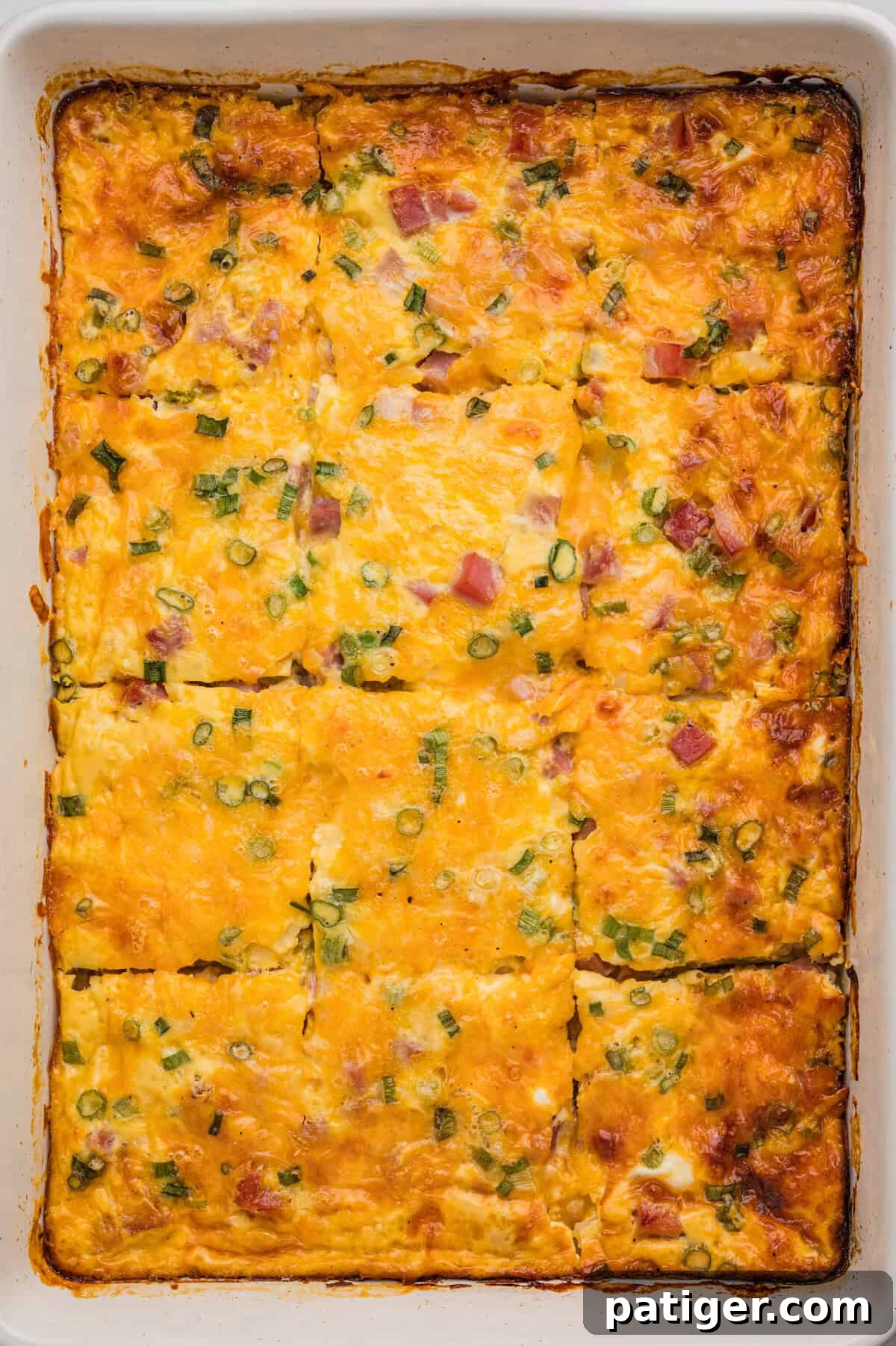 A fully baked farmer's casserole in a white dish, cut into rectangular slices with golden melted cheese, diced ham, and green onion visible on top.